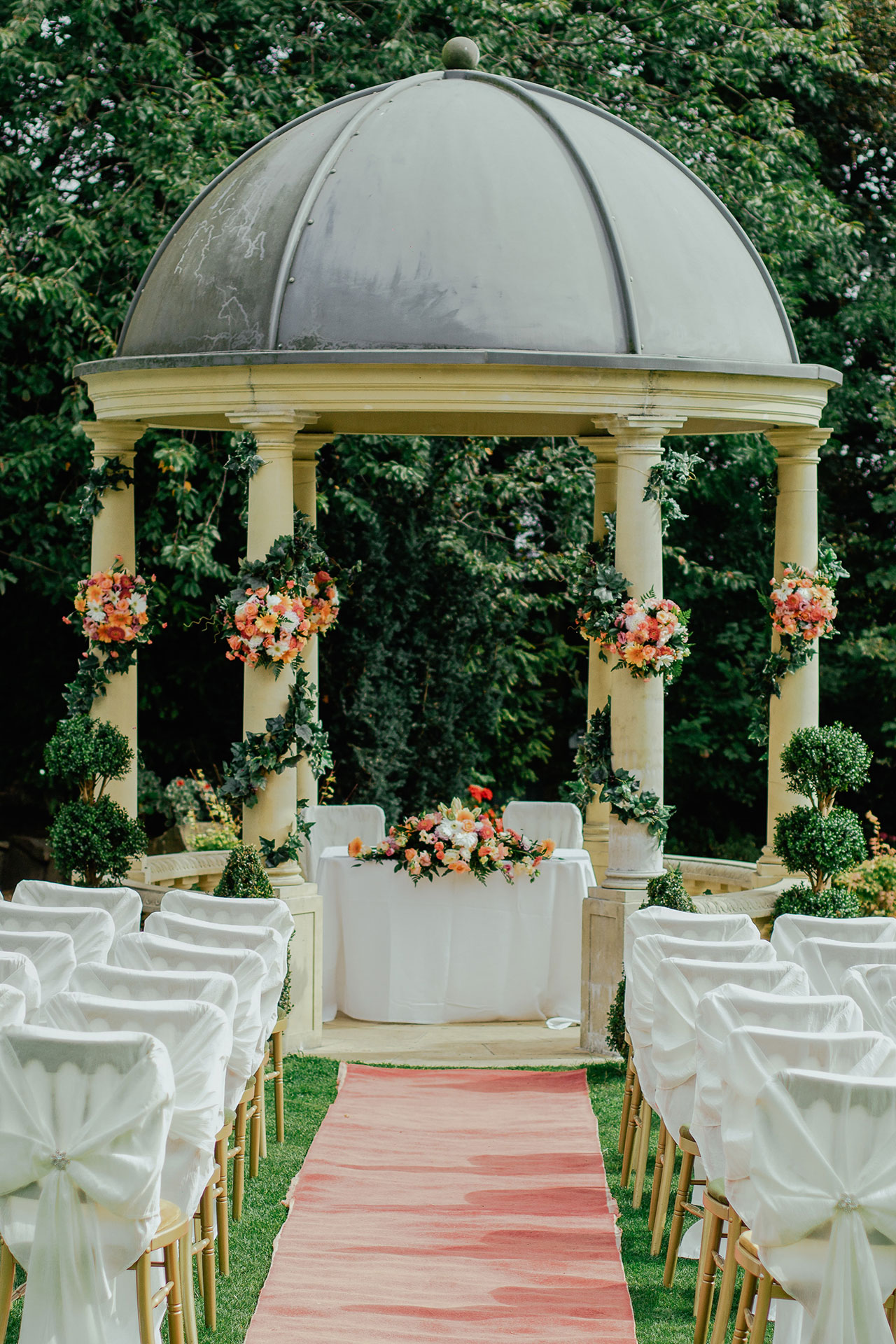 Vue en perspective des chaises de part et d'autre d'un tapis qui mènera les fiancés vers la pergola pour célébrer le mariage.